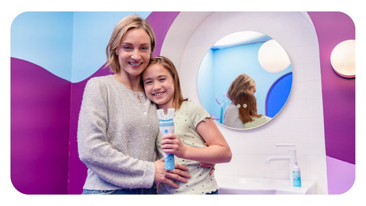 Mother hugging smiling young girl holding a light blue Willo automated kids toothbrush in a colorful bathroom setting.