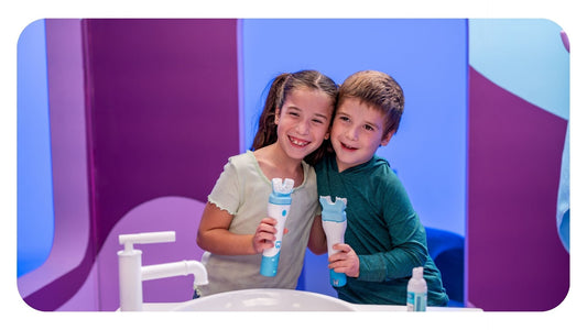 Two smiling young siblings standing together by a bathroom sink holding Willo automated kids toothbrushes.
