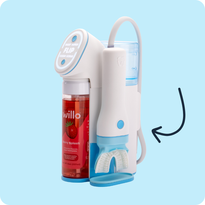 A studio product shot of the white and blue Willo automated smart toothbrush system against a light blue background. The base station setup shows an illuminated top control dial interface, a loaded red "Berry Splash" liquid toothpaste cartridge with a strawberry illustration, and the docked handheld toothbrush wand with its attached silicone mouthpiece. A dark drawn arrow graphic on the right points directly to the flexible white delivery tube connecting the wand to the base.