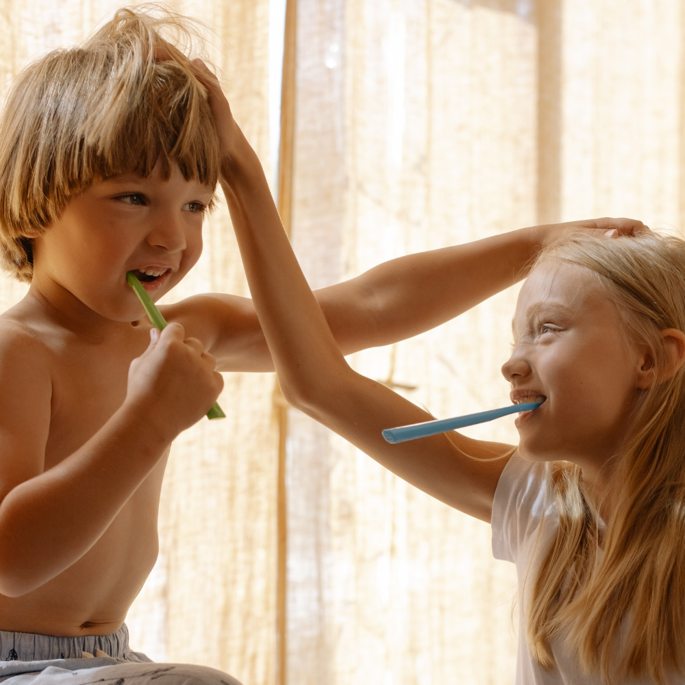 Two young children playfully brushing their teeth together with a green and blue toothbrush, smiling and interacting against a warm curtain background.
