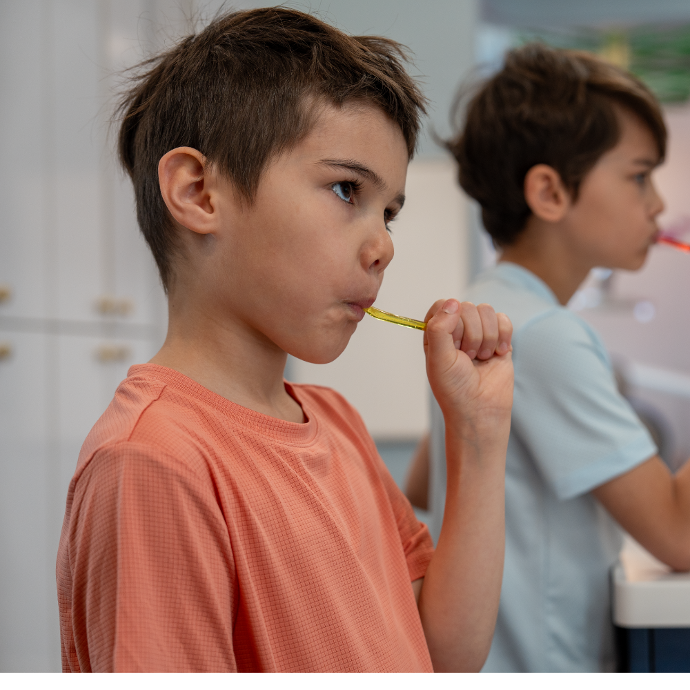 Two young boys brushing their teeth side-by-side in a bathroom, with the boy in the foreground using a yellow toothbrush.