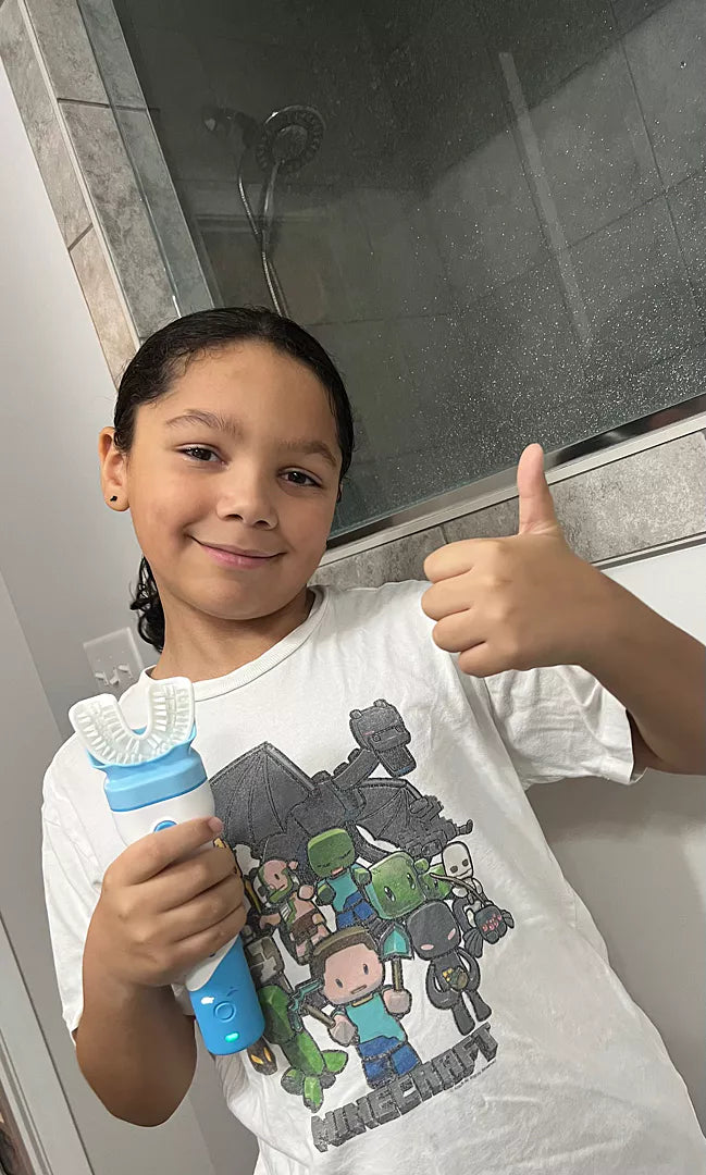 A smiling young child wearing a white Minecraft t-shirt standing in a bathroom, giving a thumbs-up while holding a white and blue Willo automated mouthpiece toothbrush. 