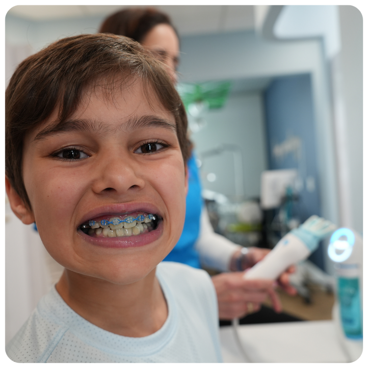 A young boy smiling to show his dental braces with blue bands during a clinical demonstration with a dentist holding a U-shaped automated smart toothbrush.