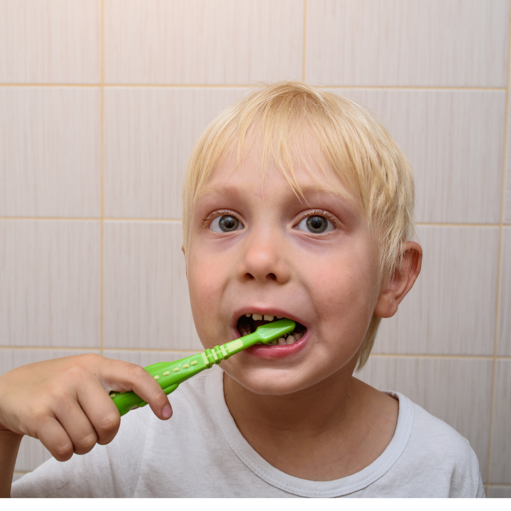 A blonde young boy looking forward while brushing his teeth with a bright green toothbrush against a light-tiled bathroom wall.