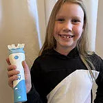 A smiling young girl with long blonde hair holding up a white and blue Willo automated mouthpiece toothbrush.