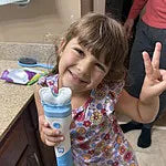 A smiling young girl with brown hair and bangs wearing floral pajamas, standing in a bathroom while holding a white and blue Willo automated mouthpiece toothbrush and flashing a peace sign.