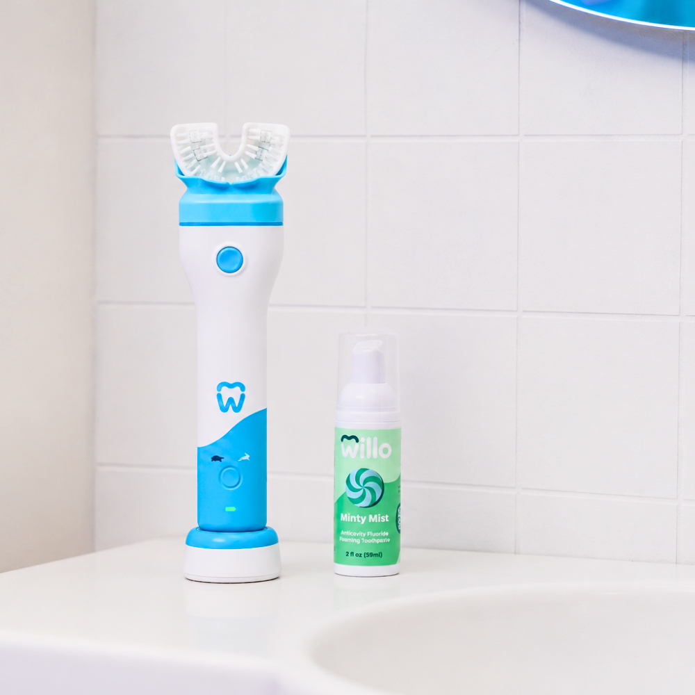 A white and blue electric toothbrush with a charging base stands next to a small green and white foaming toothpaste bottle on a white bathroom sink against a white tiled wall.